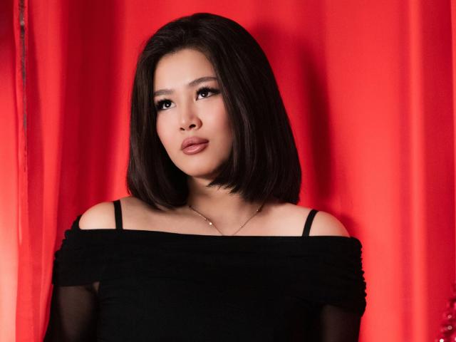 ArianaFlower, an Asian woman with a medium skin tone, has straight, shoulder-length black hair. She wears a black off-shoulder top with thin straps, revealing a small silver necklace. Her lips are slightly parted, and she has a neutral expression. The red background contrasts with her dark top and hair, highlighting her facial features. The image focuses on her upper body, with a soft light illuminating her face. Her makeup includes dark eyeliner and subtle eyeshadow.