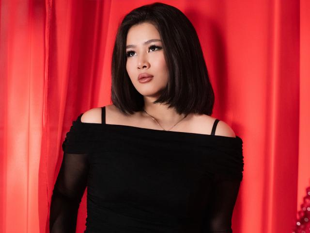 ArianaFlower with black bob, wearing off-shoulder black top, exposing black straps, red curtain background, serious expression, subtle makeup, slim build, slight chest, medium skin tone, casual pose.