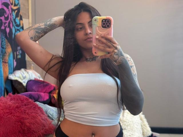 CoriPaige with long brown hair, tattoos on arms and chest, wearing a white strapless top, holding a pink phone, showing her boobs, taking a mirror selfie.