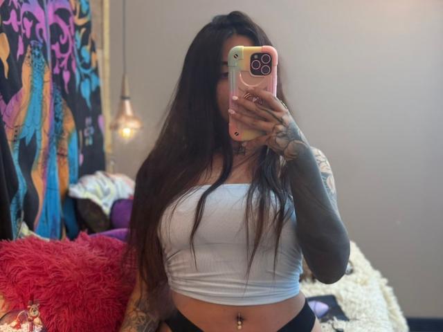 CoriPaige with long dark hair, tattoos, white crop top, black bottoms, holding pink phone, showing her tits, belly button piercing, and red fluffy pillow in the background.