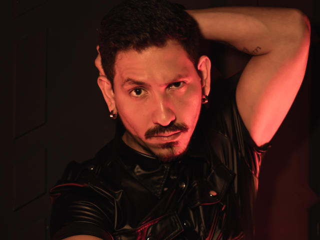 DavidBlake with a mustache, short curly hair, black leather shirt, and ear piercings, looking seductively at the camera with a red light on his right cheek.