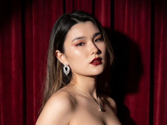AnneTLuis has a light skin tone, straight dark brown hair, and red lipstick. She wears a silver necklace with a small pendant and large, intricate earrings. Her bare shoulders are visible. The red velvet curtain in the background contrasts with her skin and hair. The lighting casts a shadow on her right side, highlighting her facial features. The image has a high contrast and a formal, almost theatrical composition.