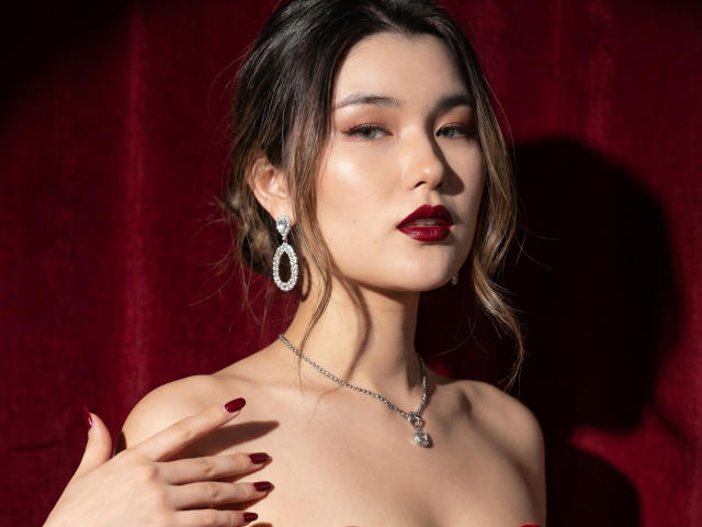 AnneTLuis with dark red lipstick, diamond earrings, and necklace, touching her bare shoulder, showing off her shiny skin and red nail polish. She's got a sexy, confident look.