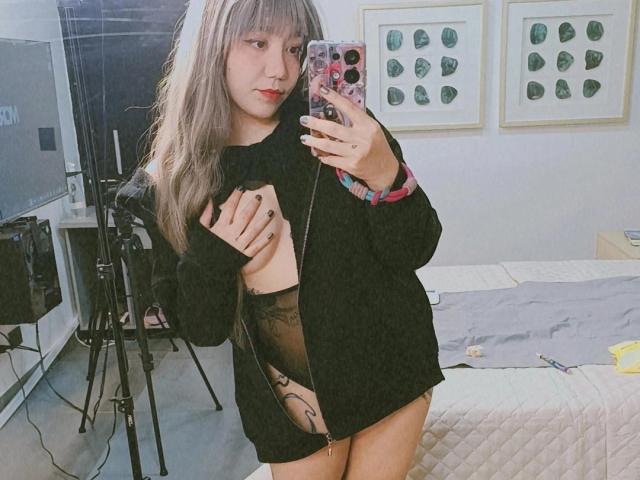 SukiBelle with long grayish-blonde hair, black long-sleeve shirt, showing one breast, tattoo on right thigh, holding phone, standing in bedroom, mirror selfie, visible bed.