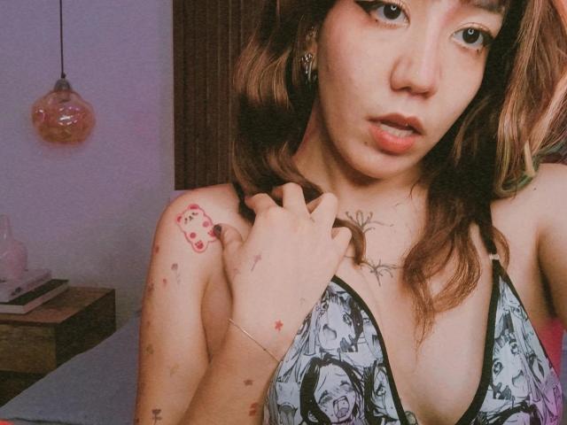 SukiBelle with wavy brown hair, wearing a black and white patterned top, has tattoos on her shoulder and chest, and is touching her collarbone with a hand. Boobs visible.
