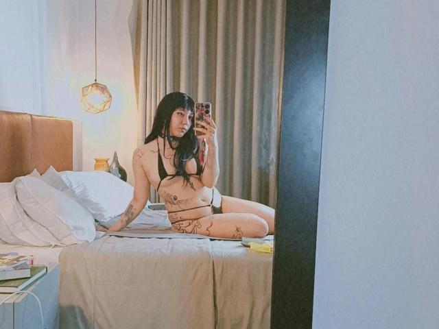 SukiBelle with long black hair, sitting on a bed, wearing a revealing black bra and thong, showing tattoos, holding a phone, reflecting in a mirror, showing her tits.