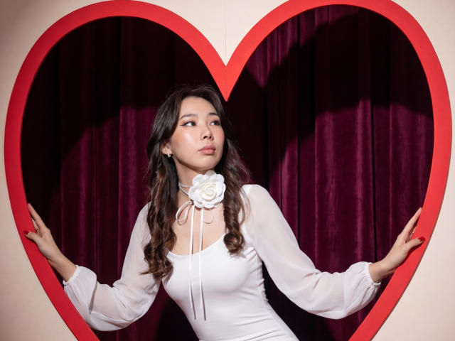AnnaReyla with wavy dark hair, wearing a white dress with a rose brooch, standing in a red heart frame, hands on heart, looking to the side, cute and sexy.