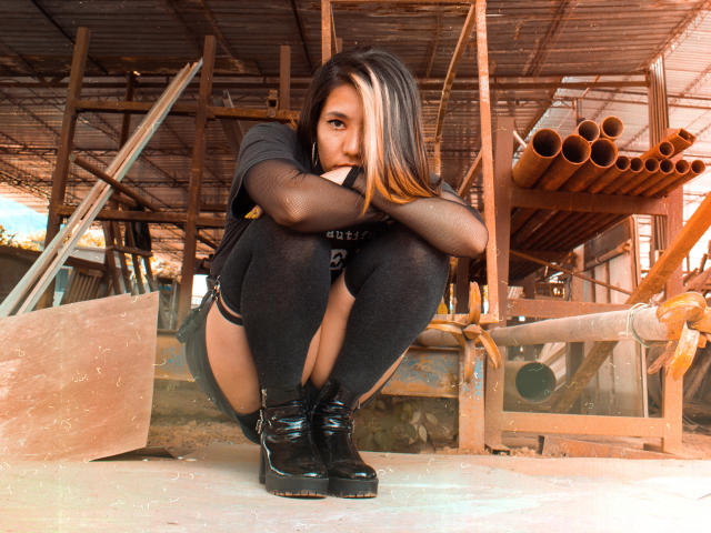 SukiBelle squatting, black shirt, black shorts, sheer sleeves, black thigh-highs, black boots, long black and blonde hair, serious expression, metal and wooden background, hands near face, crouched posture.