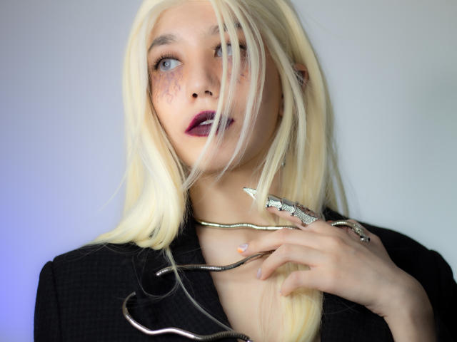 SukiBelle with blonde hair, purple lipstick, freckles, black jacket, and a silver snake necklace, holding it near her chest, looking to the side with a slightly open mouth.