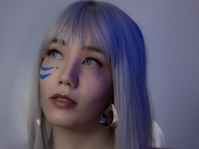 SukiBelle has pale blue eyes, white hair with bangs, and blue face paint on the left cheek. She wears metallic ear cuffs and gazes upward with a slight smile.
