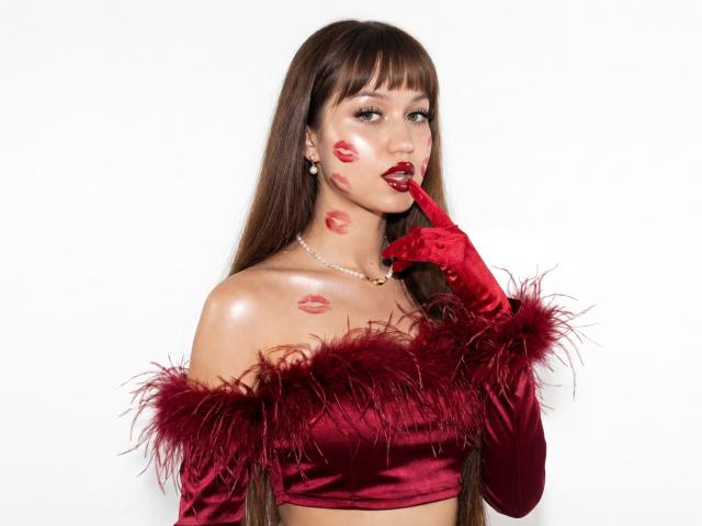 TiffanySweetex has long brown hair with bangs, wearing a red off-shoulder velvet top with fluffy feather trim and matching red gloves. Her face has red lipstick marks, and she's biting her finger with a seductive expression. She's wearing a pearl necklace. The background is plain white, making TiffanySweetex the clear focus. The red velvet and feathers contrast with her fair skin. The image is sharp with a modern, sexy vibe.