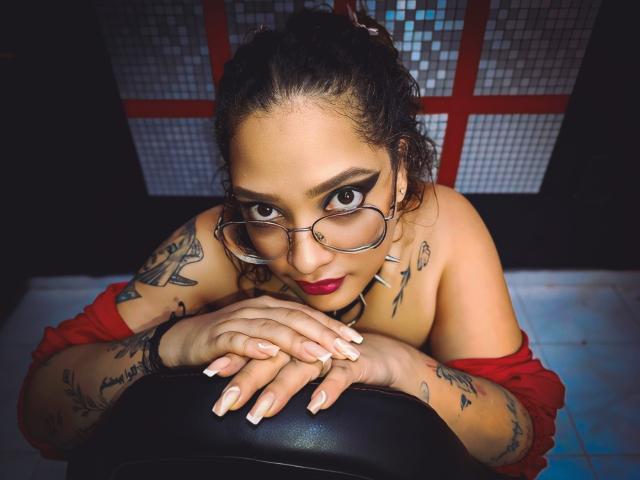 DanielaPatherson with tattoos, glasses, red shirt off, hands on black chair, looking up with dark eyes, red lipstick, and nails, has several tattoos, and a nose ring.
