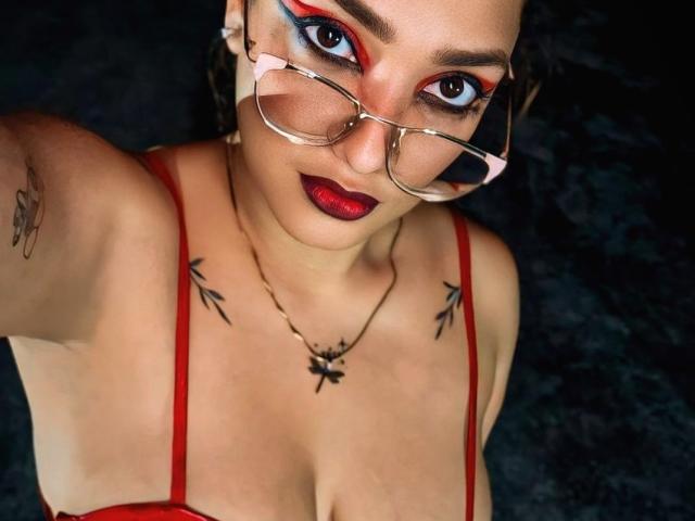 DanielaPatherson with red lipstick, glasses, and red bra, showing tits with tattoos on chest, and arms, looking up with dark, bold makeup, in a dark setting.