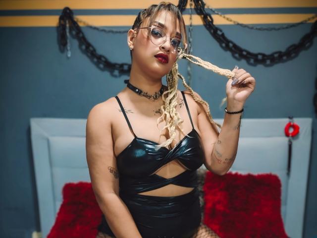 DanielaPatherson with blonde dreadlocks, wearing glasses, black leather crop top, choker, holding her hair, red pillows, tattoos, and a black ring on her right hand.