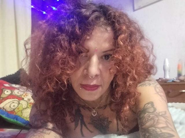 MariPosatatto with curly red hair, nose ring, lip piercing, and tattoos, looking at the camera with red lipstick, lying on a bed, visible chest and arms.