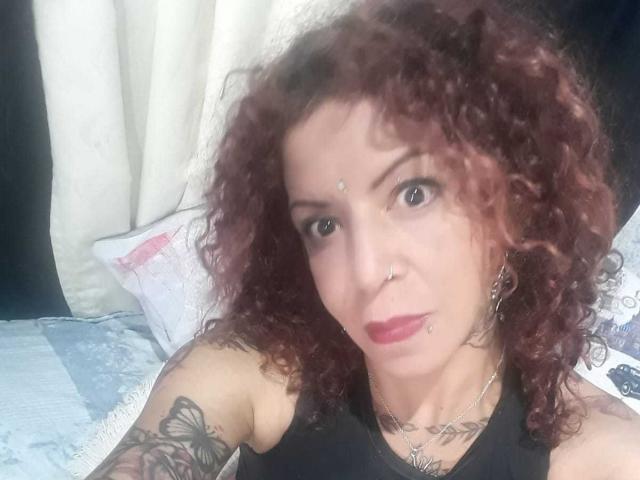 MariPosatatto has curly red hair, a nose ring, and lip piercing. She's wearing a black top, showing a butterfly tattoo on her right arm. Her tits are small.