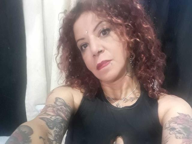 MariPosatatto with curly red hair, nose ring, and tattoos on arms, wearing black cut-out top, showing small tits, and pink lipstick, taking a selfie.