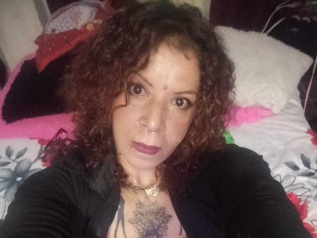 MariPosatatto with curly brown hair, nose ring, and dark lipstick, wearing a black shirt, showing a tattoo on her chest, looking at the camera, lying on a bed.