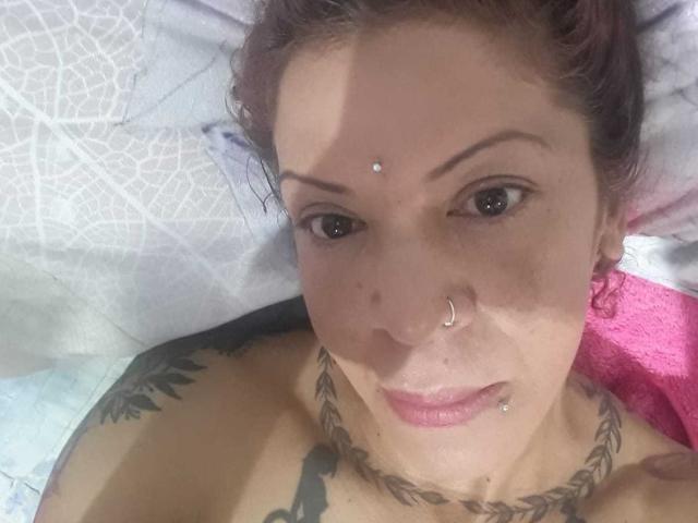 MariPosatatto, topless with tattoos, has a nose ring, lip piercing, and facial piercing, lying on a bed, showing off her tits and a necklace. Her pink towel is visible.