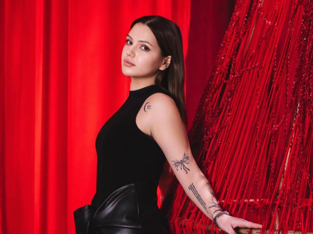 SophiaGrant with long brown hair, black sleeveless top, and black leather pants, showing tattoos on her arms, standing against red string decor, looking at camera with a slight smile.