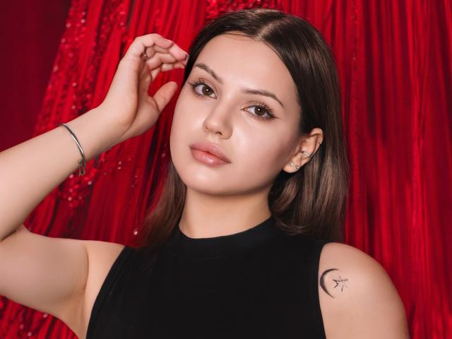 SophiaGrant has brown eyes, medium-length brown hair, and a black sleeveless top. She's touching her hair with a silver bracelet, showing a crescent tattoo on her right arm.