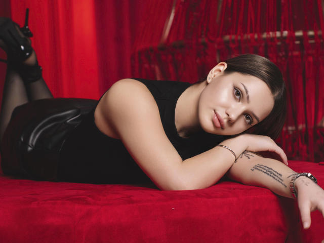 SophiaGrant, lying on red velvet, wearing black sleeveless top, black leather shorts, black heels, tattoo on right forearm, brown hair, looking at camera, arms resting on red bed.