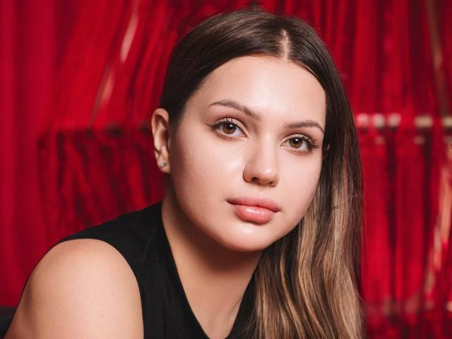 SophiaGrant has smooth, fair skin, brown eyes, and long brown hair. She's wearing a black sleeveless top and has small, subtle earrings. Her lips are slightly pouty.