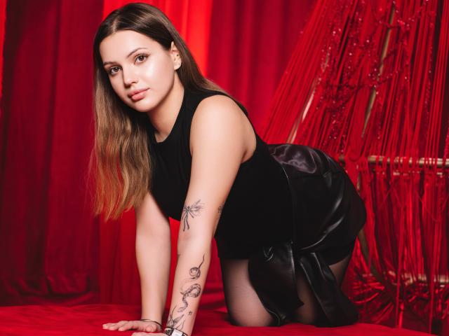 SophiaGrant on all fours, wearing a black sleeveless top and black leather skirt, with tattoos on her arms, in a red-themed setting, looking directly at the camera.