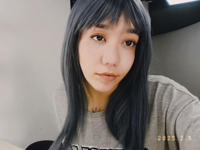 SukiBelle with long black hair, tear on left cheek, small tattoo below chin, wearing grey shirt, looking at camera, 2025.5 date stamp. Slight pout, subtle makeup, soft expression.