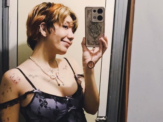 SukiBelle with short, blonde and brown hair, smiling, holding phone, wearing black lace top, pearl necklace, tattoos, taking mirror selfie, showing her tits and a cute face.