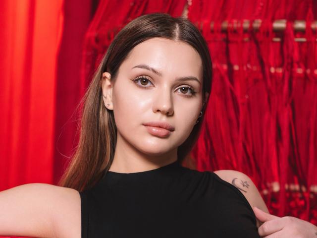 SophiaGrant with smooth, shiny skin, brown eyes, and subtle makeup, wearing a black sleeveless top, showing a small tattoo on her right shoulder, against a red backdrop.