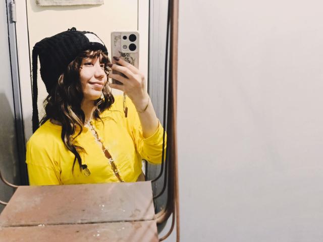 SukiBelle with wavy brown hair, black knit beanie, yellow shirt, and brown necklace, taking a mirror selfie with a subtle smile, holding a phone with a white case.