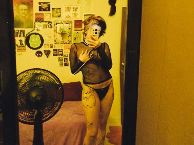 SukiBelle with short brown hair, wearing a sheer black top and black panties, holding a phone in a mirror selfie. Her tits are small, and she has a tattoo on her thigh.