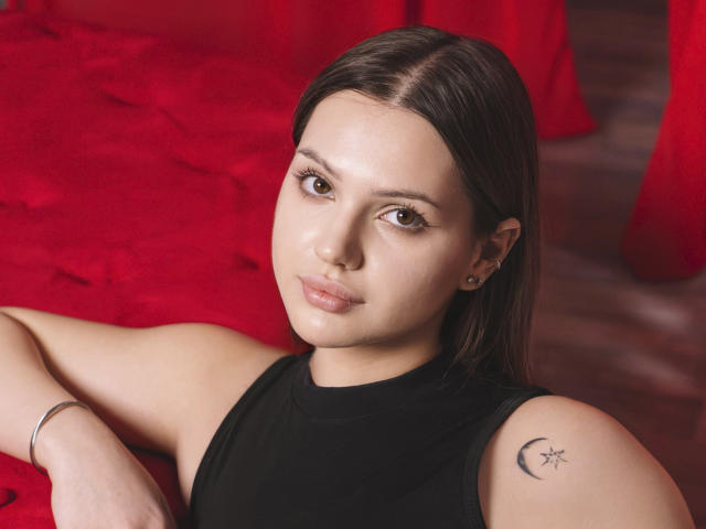 SophiaGrant has light skin, brown eyes, and straight dark brown hair, wearing a black sleeveless top, with a crescent star tattoo on her right shoulder, and a silver bracelet.