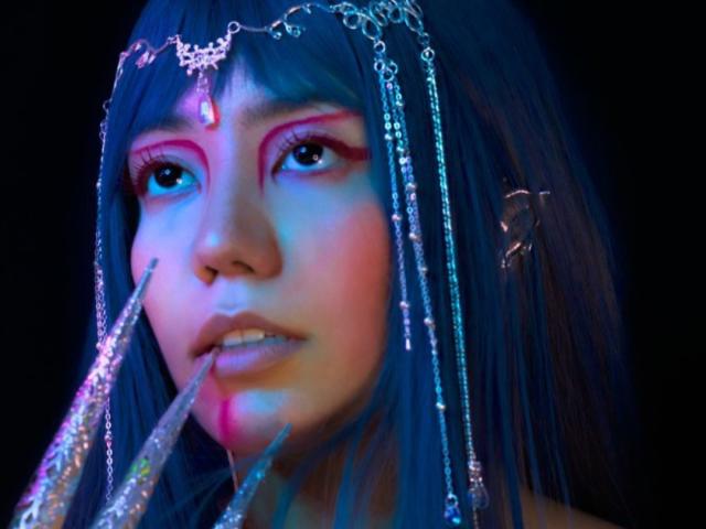 SukiBelle with blue hair, wearing a metal chain headpiece, neon pink eye makeup, and glittery nails, touching her lower lip. Glittery, metal chains, and neon colors.