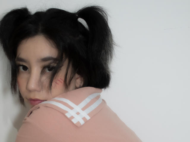 SukiBelle with black puffy twin-tails, light pink top, white criss-cross straps, slight red makeup on cheek, looking over shoulder, white background.