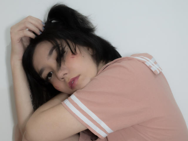 SukiBelle with messy black hair, pink shirt with white stripes, arms crossed, looking at camera with slightly pouty lips, small red scratch on right cheek.