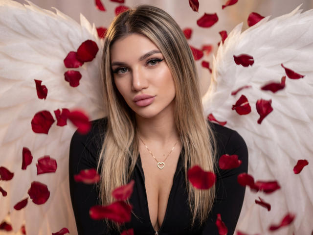 SelenaeMoore with white angel wings, red rose petals, and a heart necklace, wearing a black top, showing her boobs, with blonde hair and a neutral expression.