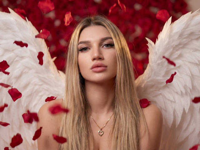 SelenaeMoore has long, straight blonde hair and a neutral expression. She's centered with white, feathery angel wings. Red rose petals float around her. She wears a gold heart necklace. The background is a blurred, dense red rose. The image has a soft, romantic vibe with a focus on SelenaeMoore's calm, confident presence. The white wings contrast sharply with the red petals and background. The rose petals add a sense of movement. The overall composition is balanced and elegant.