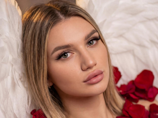 Close-up of SelenaeMoore with light brown, straight hair and green eyes. She has a fair complexion, full lips with a subtle pink gloss, and thick, dark eyebrows. She's looking slightly to the right. Her makeup is subtle with long, thick eyelashes. She's wearing a red rose-petal garment. White feather-like textures are in the background. The image has a soft, warm light. SelenaeMoore's expression is calm and slightly pensive. The composition focuses on her face and upper chest.