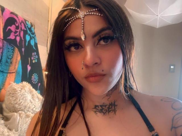 CoriPaige heeft bruin haar, een neusring en een hoofdband met een ketting. Ze heeft tatoeages, dikke eyeliner en een ketting om. Haar borsten zijn zichtbaar, en ze kijkt naar de camera..