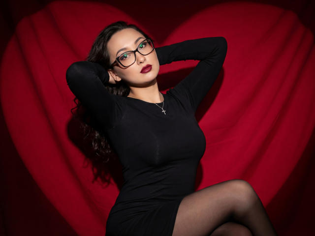 EvaSarazardje, a woman with fair skin and long, curly dark hair, wears black-rimmed glasses and a tight black long-sleeve dress that accentuates her medium-sized breasts. She sits with her arms raised, hands behind her head, and dark red lipstick. She has a silver cross necklace and black tights. The background is a large red heart, creating a dramatic contrast with her black outfit. The image is sharply focused, with a slightly shadowed effect highlighting her facial features and upper body.