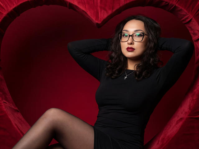 EvaSarazardje with glasses, black dress, and cross necklace, hands behind head, red heart background, black pantyhose, sitting, sexy, dark wavy hair, red lipstick.