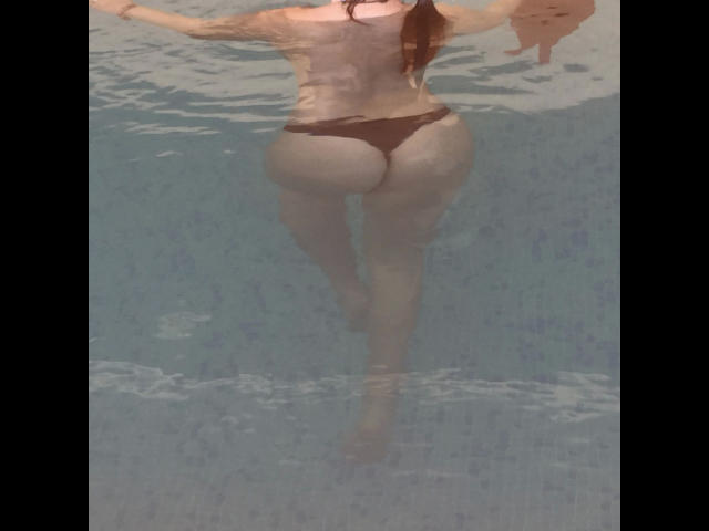 SweetSonia-hot's back is to the camera, wearing dark brown thong underwear, with her bare ass and legs visible in a pool, her arms out, and hair in a ponytail.