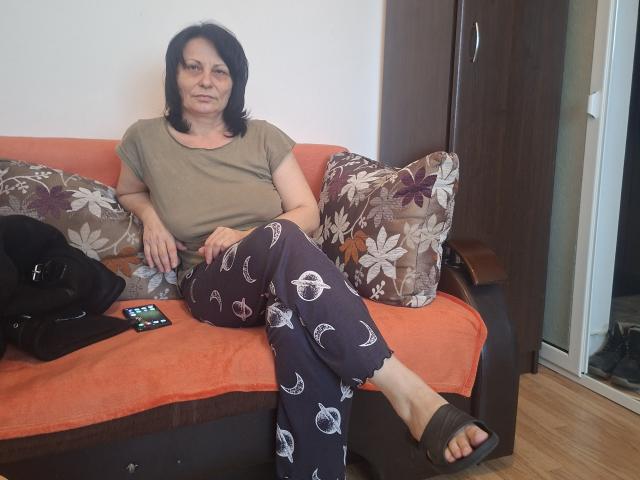 Jessyca6 is sitting on an orange sofa with floral and geometric patterned pillows. She has medium-length black hair, a serious expression, and is wearing a brown t-shirt and dark blue pajama pants with white moon and eye patterns. Her right arm rests on the sofa, while her left hand is on her knee. She's wearing black sandals. A black bag and a remote control are on the sofa to her left. The background includes a wooden wardrobe and a glass door.
