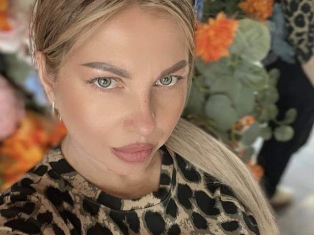 A close-up of AmandaSexyX with light skin and blonde hair in a ponytail. She has green eyes, dark eyebrows, and pink lipstick. She wears a leopard-print top. Her makeup is subtle with a natural look. The background is blurry with orange flowers and green leaves. Her expression is neutral. The image is sharp with a focus on her face. She has small, gold, hoop earrings. The lighting is even.