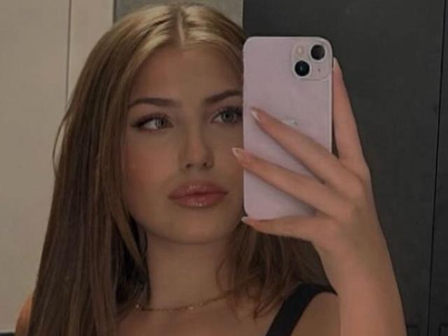 KattyLips with blonde hair, holding a pink phone, showing her face with full lips and black top, looking into the camera with a neutral expression.