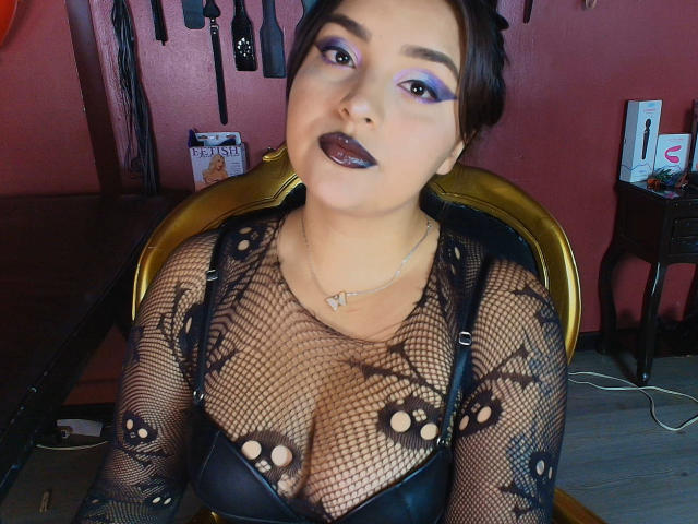 MissSubmissive mit dunklem Lippenstift, lila Lidschatten und einem schwarzen Netzoberteil, das ihre Brüste zeigt, trägt einen schwarzen BH und sitzt auf einem goldenen Stuhl in einem Raum mit BDSM-Ausrüstung..