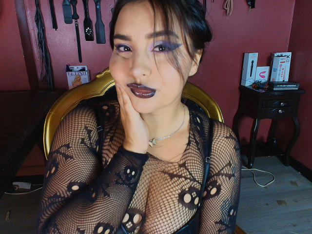 MissSubmissive mit dunklem Lippenstift und lila Lidschatten, trägt ein schwarzes Netzoberteil, das ihre Brüste zeigt, und hat ihre Hand im Gesicht. Sie sitzt in einem Raum mit BDSM-Ausrüstung..