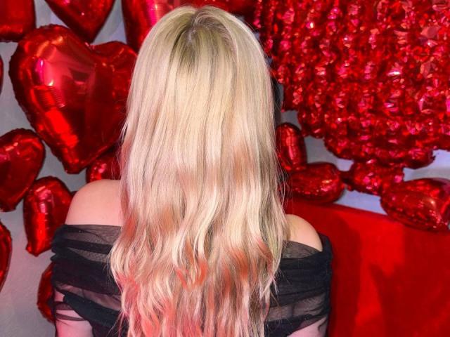 CrystalHope's blonde, wavy hair, off-shoulder black dress, back to camera, surrounded by shiny red heart balloons and a large red heart decoration.